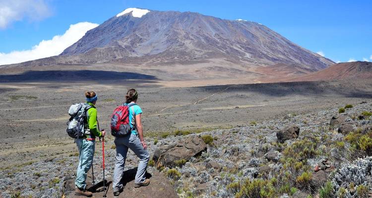Wandelaars staan op een rotsachtige uitstekende rots en kijken uit over het uitgestrekte Shira Plateau en de top van de Kilimanjaro eromheen.