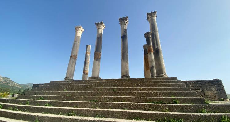 Zes rechtopstaande Romeinse zuilen bovenop brede stenen trappen onder een strakblauw hemel.