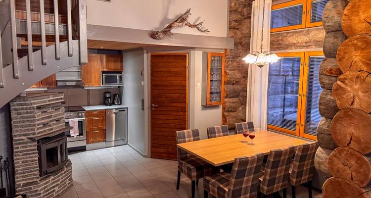 Intérieur spacieux de chalet alpin avec foyer en pierre, cuisine en bois et chaises de salle à manger à carreaux sous une décoration de bois de cerf.