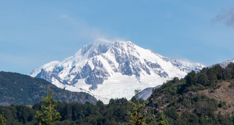 Isolated snow-covered peak rises above forested foothills under a clear sky.