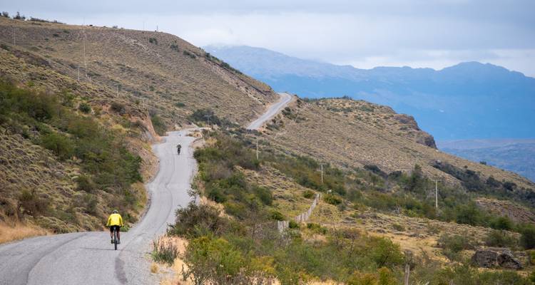 Cyclist ascends a remote paved road along arid mountain slopes with switchbacks ahead.