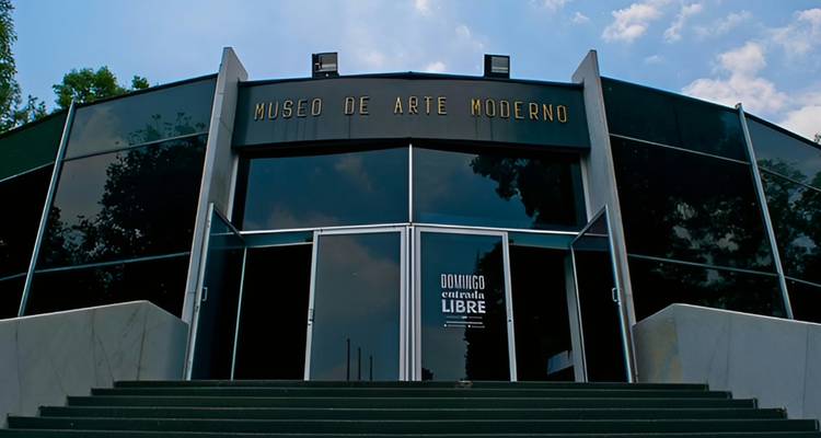 Frontfassade des Museo de Arte Moderno mit großen Glaspaneelen und zweisprachiger Beschilderung, fotografiert von der darunter liegenden Treppe aus.