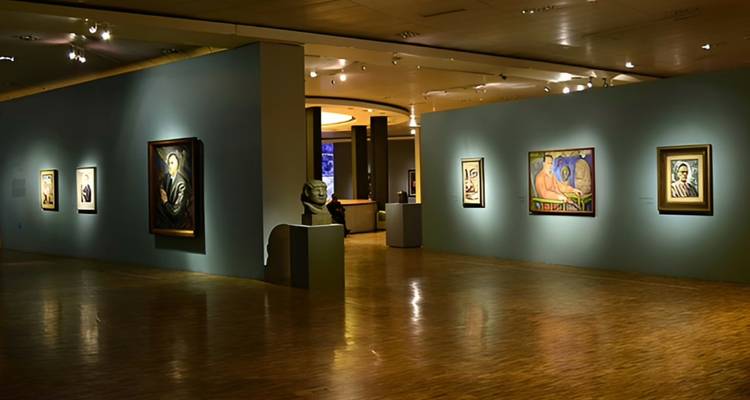 Schwach beleuchteter Kunstgalerie-Saal mit gerahmten modernen Gemälden an blaugrünen Wänden und polierten Holzböden.