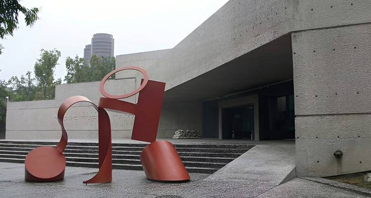 Modernes Betonmuseumsgebäude mit einer großen roten abstrakten Außenskulptur neben breiten Treppen.