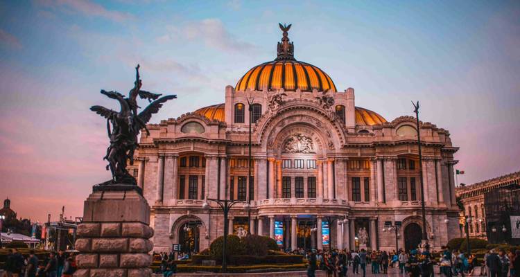 Der Palacio de Bellas Artes erleuchtet sich in der Abenddämmerung unter einem pastellfarbenen Himmel, mit Menschenmengen auf dem Platz und einer geflügelten Statue im Vordergrund.