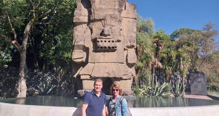 Zwei Reisende posieren vor der riesigen Basaltstatue eines Regengottes mit Springbrunnen auf einem üppigen Außenplatz.