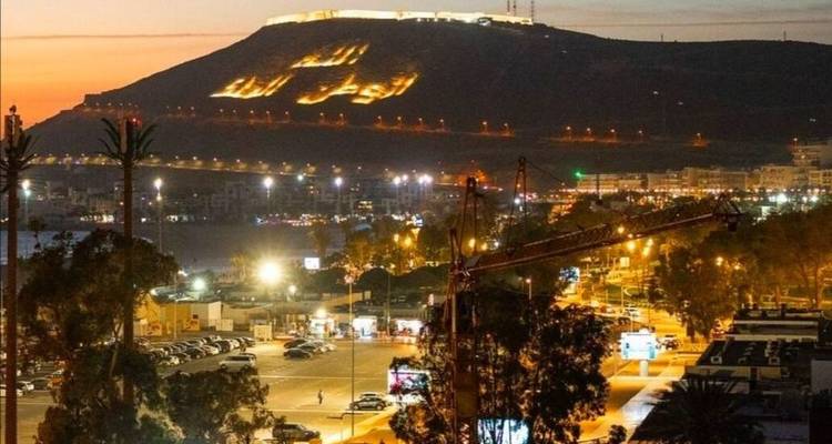 Paysage urbain d'Agadir au crépuscule avec l'inscription illuminée sur la colline et les lumières du port scintillant en bas.