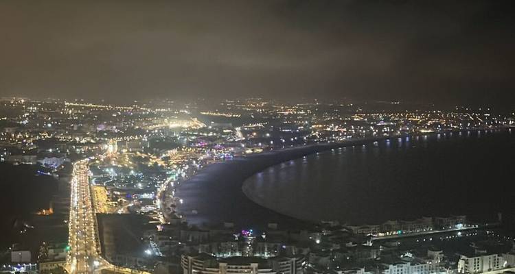 Vue aérienne nocturne de la baie incurvée d'Agadir bordée de lumières urbaines sous un ciel nuageux.
