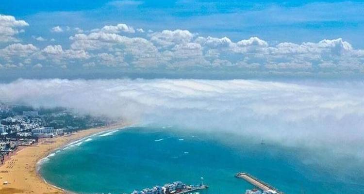 Vue aérienne diurne d'une baie turquoise partiellement voilée par un banc de brouillard marin avec plage de sable et port en contrebas.