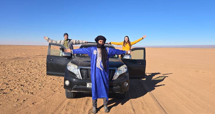 Turistas con los brazos extendidos se paran frente a un vehículo 4x4 en el vasto desierto bajo un cielo azul despejado.
