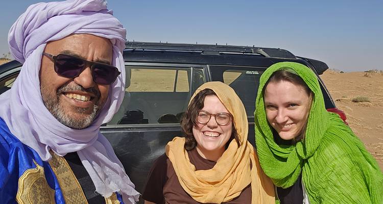 Guía y dos viajeros con pañuelos de colores sonriendo junto a un vehículo en el desierto.