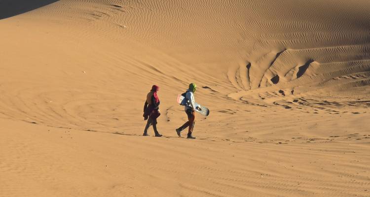 Dos personas caminando sobre arena ondulada llevando una tabla de snowboard para hacer sandboarding en una duna del desierto.
