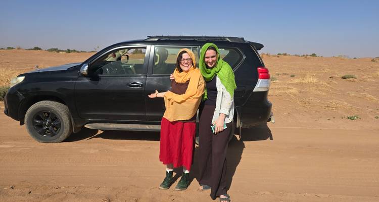 Dos mujeres con ropa de viaje de colores brillantes posan junto a un vehículo 4x4 negro en una pista de arena del desierto.
