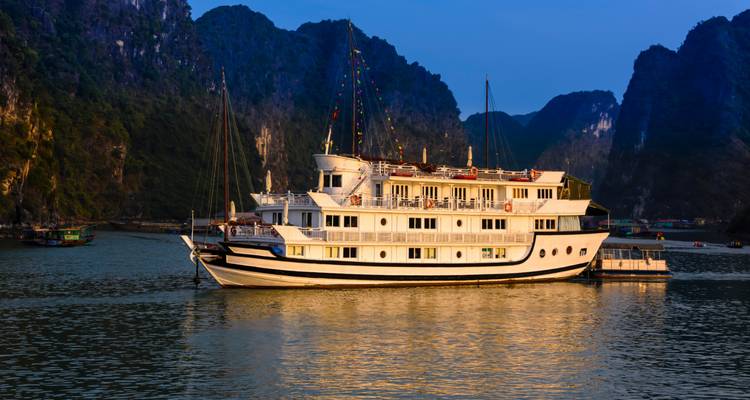 Elegant wit cruiseschip verlicht door warm avondlicht, voor anker tussen donkere kalksteen toppen.