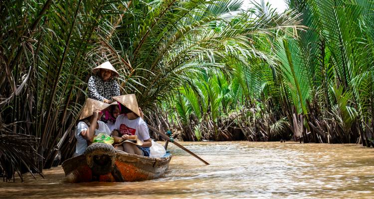 Toeristen met puntige hoeden die met een houten boot door dichte palmkanalen in de Mekong peddelen.