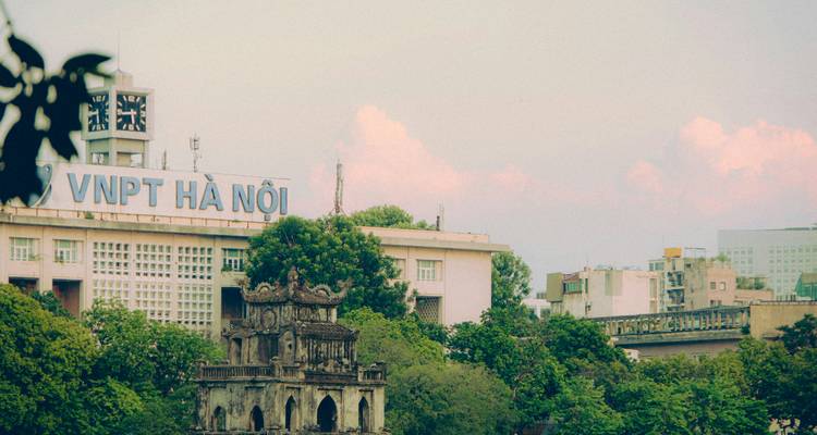 Stadsscène van Hanoi met de Schildpaddentoren op het Hoan Kiem-meer omlijst door bomen en een groot VNPT Ha Noi-gebouw in zacht avondlicht.