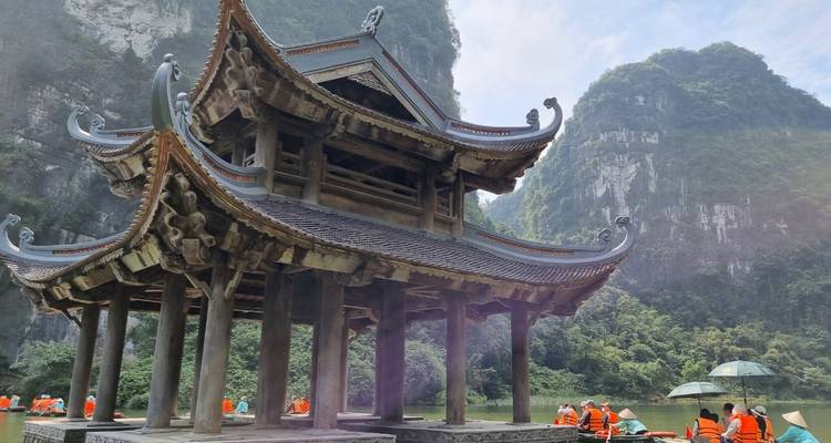 Traditionele houten Vietnamese paviljoen op palen boven het water met dramatische kalkstenen rotsen op de achtergrond en toeristen in oranje reddingsvesten in de buurt.