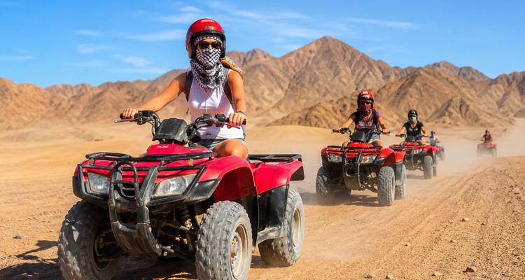 Les aventuriers chevaucent des quads rouges sur un sentier désertique brûlé par le soleil avec des montagnes escarpées au loin.
