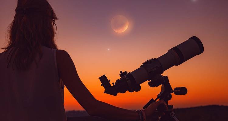 Un observateur d'étoiles en silhouette regarde à travers un télescope tandis qu'une lune croissante brille dans un ciel crépusculaire aux couleurs éclatantes.