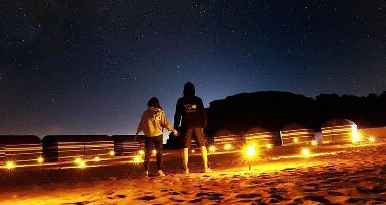 Un couple se tenant la main sous un ciel étoilé dans un campement désertique éclairé aux chandelles évoque une atmosphère romantique.