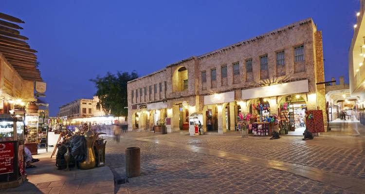 Les bâtiments en pierre traditionnels illuminés à la tombée de la nuit dans la place de marché animée du Souq Waqif de Doha, avec quelques piétons flous traversant les pavés.