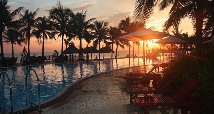 Piscine à débordement bordée de palmiers reflétant les couleurs ardentes du coucher de soleil avec des transats vides le long de la terrasse humide.