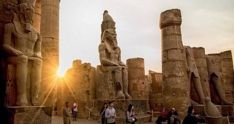 Les gigantesques statues de pharaon assis du temple de Louxor brillent sous le soleil couchant tandis que les touristes explorent les lieux en contrebas.