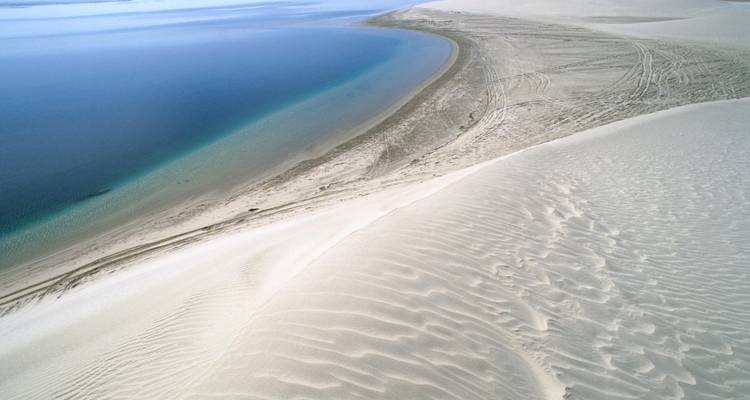 Vue panoramique depuis la crête d'une dune de sable pâle jusqu'aux eaux bleu-vert tranquilles le long de la côte.