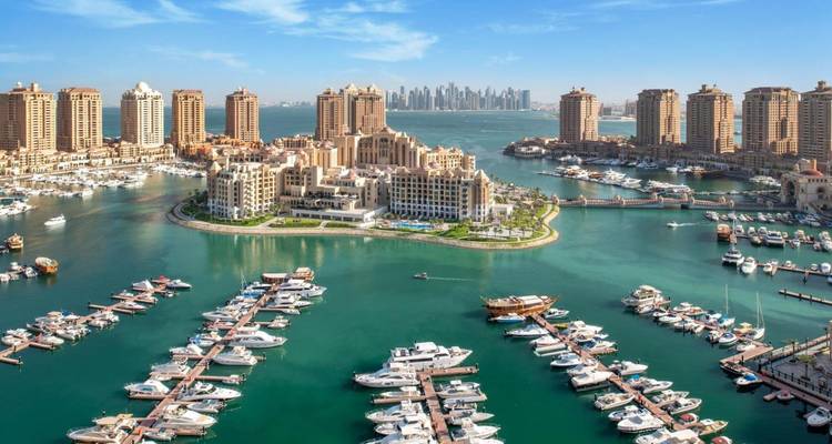 Panorama aérien de la marina de The Pearl remplie de yachts, entourée de tours de luxe avec l'horizon de Doha en arrière-plan.