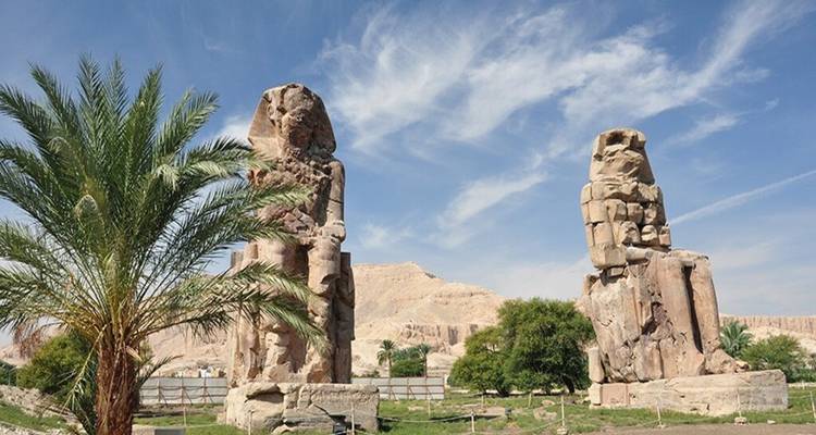 Les deux colosses de Memnon se dressent majestueusement au milieu des palmiers avec des falaises désertiques en arrière-plan.