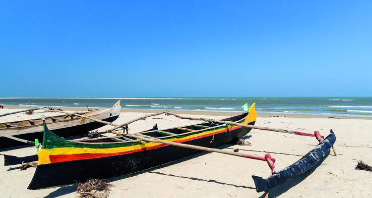 Traditional wooden outrigger canoes rest on a quiet sandy beach under a clear blue sky.