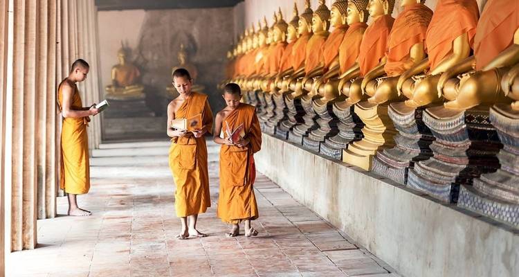 Binnengang van een tempel met gouden Boeddha-beelden aan weerszijden, waar twee jonge monniken uit boeken lezen.
