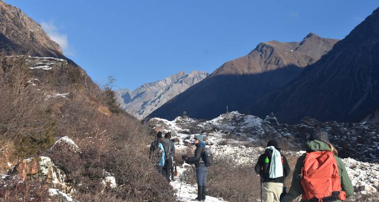 Los excursionistas avanzan por un sendero alpino nevado flanqueado por dramáticos picos marrones bajo cielos azules despejados.