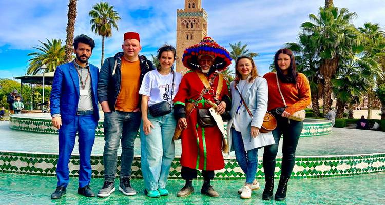 Groep bezoekers die poseren bij een tegelvoorziene fontein met de Koutoubia-moskee die erachter uittorent onder een blauwe hemel.
