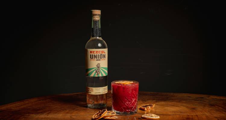 Flasche Mezcal Unión neben einem roten Cocktail und getrockneten Orangenscheiben auf einem rustikalen Holztisch.