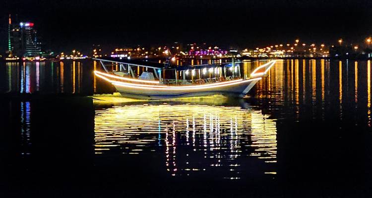 Illuminated dhow boat glides across mirror-like harbor waters at night.
