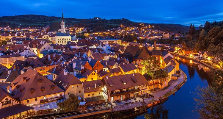 Vista nocturna sobre la curva del río y el pueblo medieval de Cesky Krumlov con tejados rojos, iluminado por luces cálidas.
