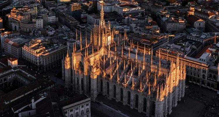Luftaufnahme der Mailänder Kathedrale in der Dämmerung, beleuchtet durch warmes Sonnenlicht inmitten der Stadtdächer.