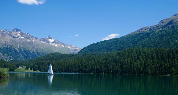 Ruhiger Alpensee mit einem einsamen weißen Segelboot, umgeben von bewaldeten Hängen und schneebedeckten Gipfeln.