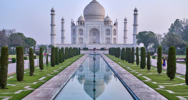 Iconisch frontaal zicht op de Taj Mahal weerspiegeld in het lange bassin bij zonsopgang met weinig bezoekers aanwezig.