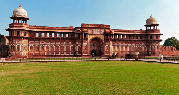 Breed uitzicht op de rode zandstenen gevel van het Agra Fort tegen een heldere blauwe lucht en een verzorgd gazon.