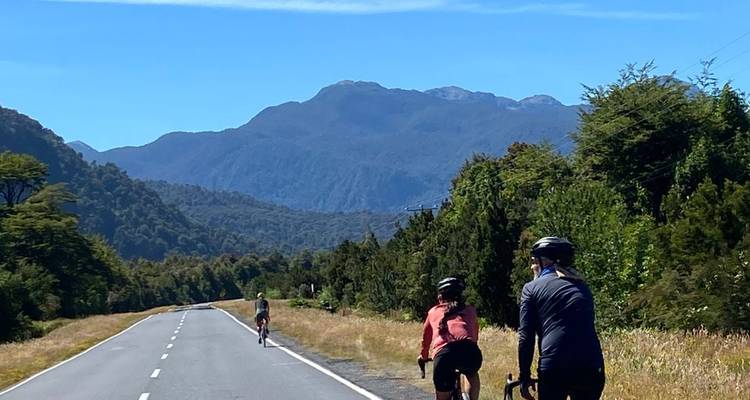 Cyclists ride along an empty paved road through lush forested valley with blue mountains in the distance