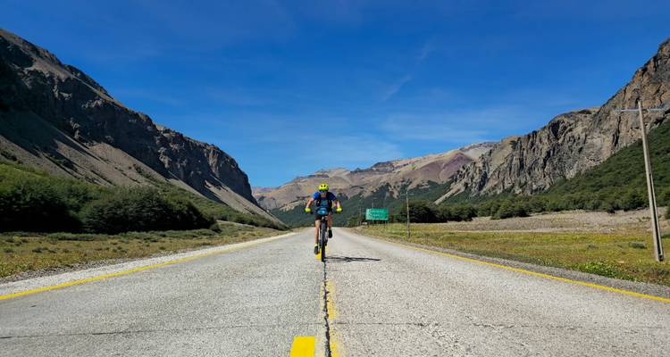 Solo cyclist pedals down the centre of a scenic mountain highway flanked by steep rocky cliffs under clear sky