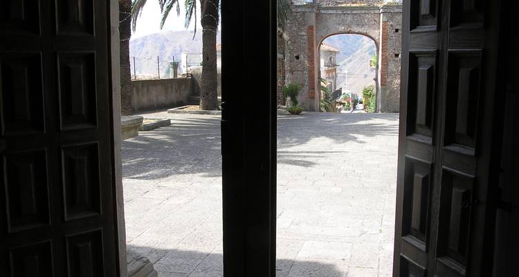 Vue ombrée depuis l'intérieur de portes en bois donnant sur une arche en pierre et une place ensoleillée.
