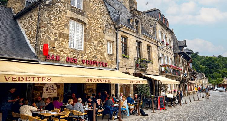 Bustling café terrace with diners beneath cream awnings on a cobblestone street lined with historic stone buildings and flower boxes.