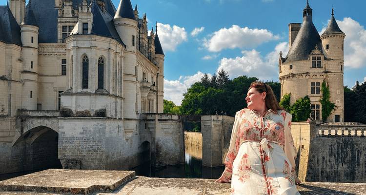 Smiling woman sitting on a stone ledge with fairytale château towers rising behind her under a blue sky.