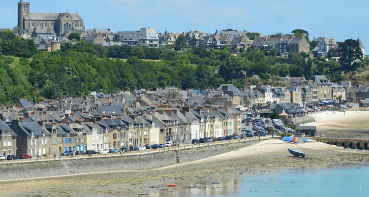 Seaside town with rows of stone houses along a sandy shore backed by green hills and a hilltop church.