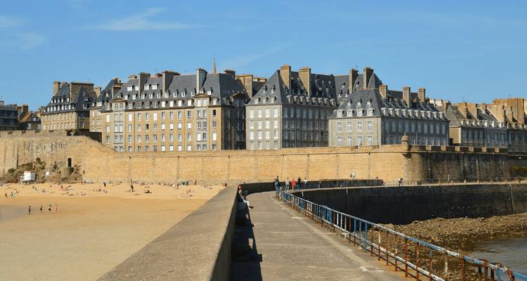 Massive stone ramparts and grey-roofed buildings of the fortified city of Saint-Malo rising above a golden beach.