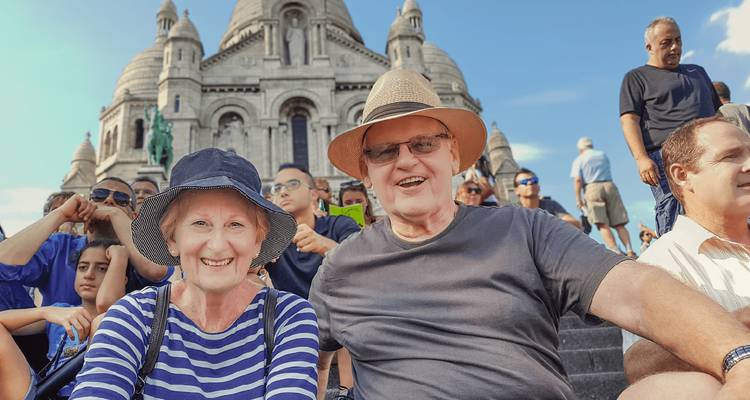 Smiling senior couple and other tourists seated on the steps in front of the Sacré-Cœur Basilica on a sunny day.