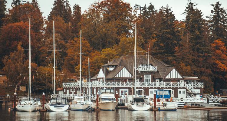 Historisches Bootshaus und Segelboote, umrahmt von Herbstbäumen auf ruhigem Wasser.